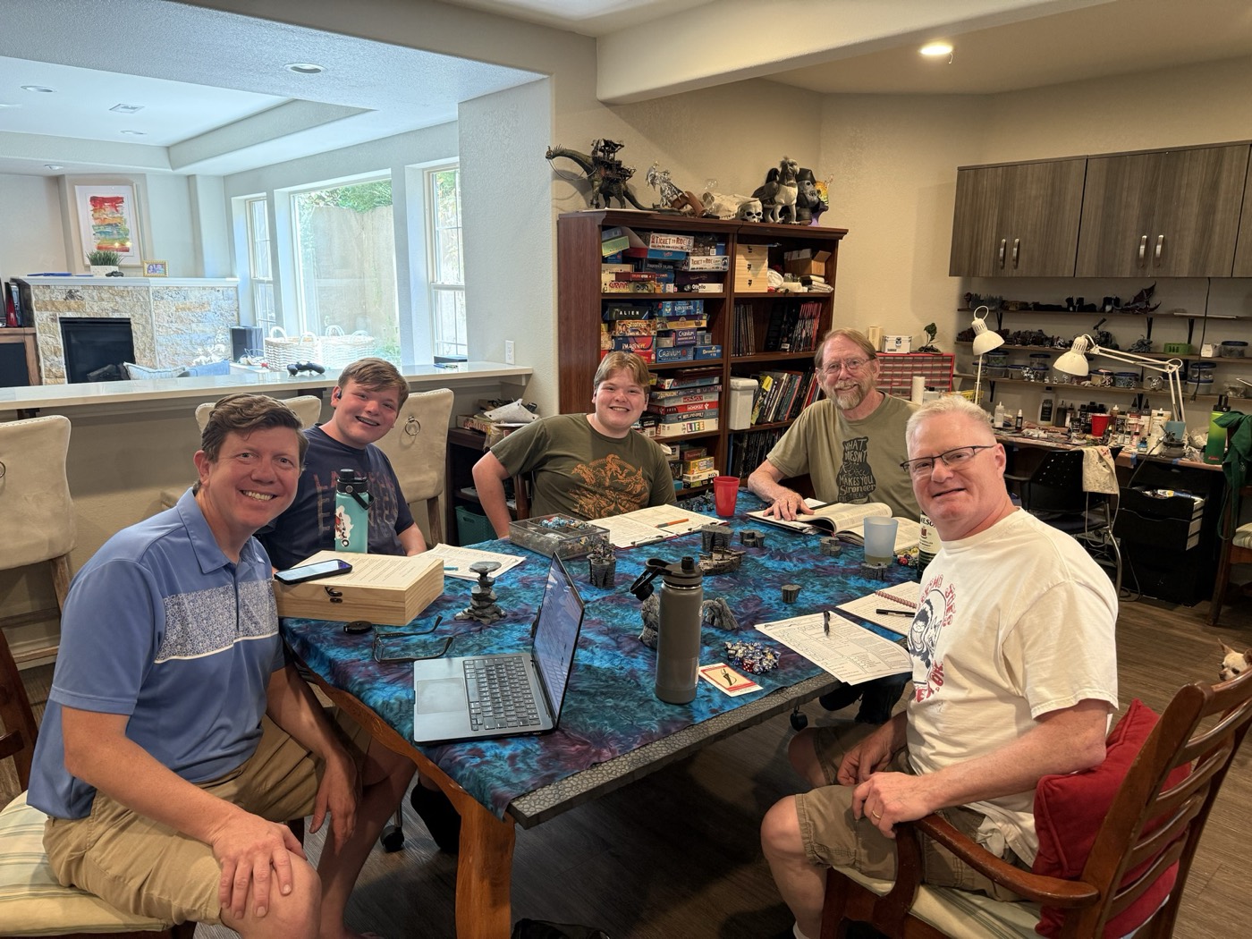 Five people around a game table at home, with a laptop, dice, character sheets and a bookcase of games behind them.