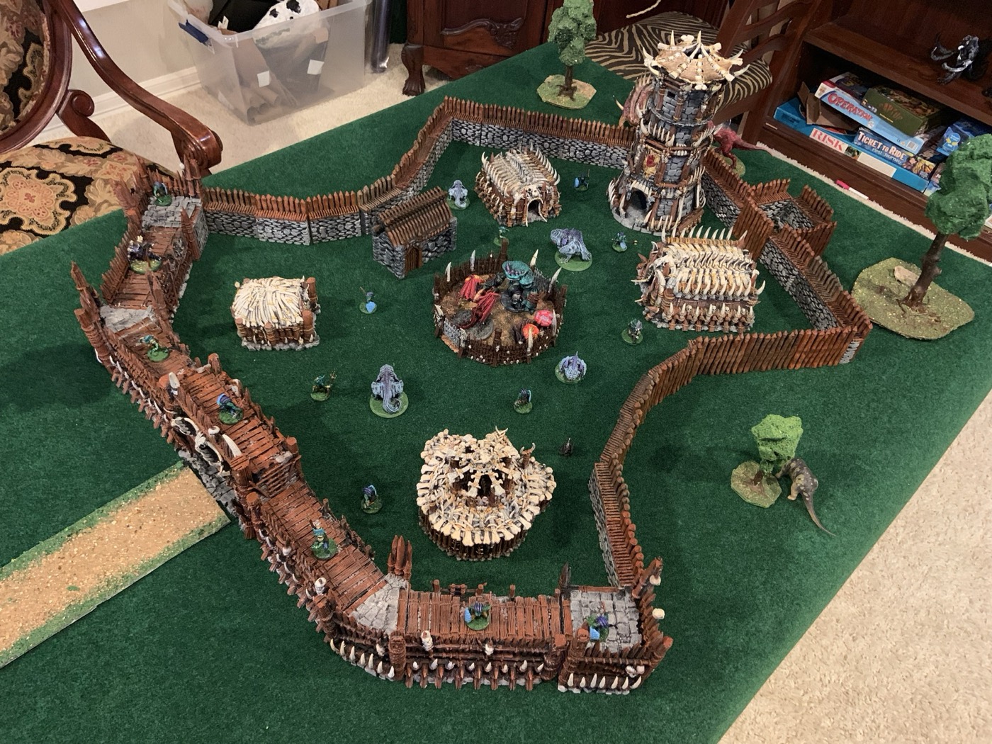 A tabletop camp seen from above: bone huts, a palisade wall, a watchtower and scattered miniatures on green felt.